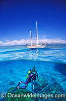 Half under and half over water picture of Scuba Diver beneath sailing yacht, exploring tropical Coral reef. Fijian Islands